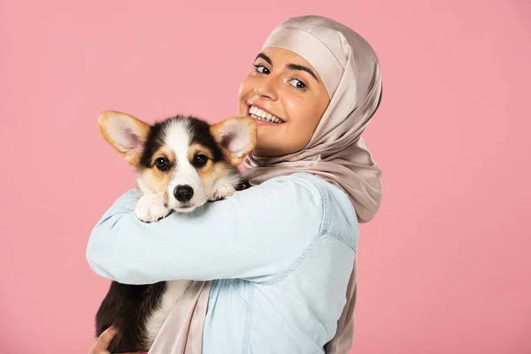 Beautiful muslim woman in hijab holding Welsh Corgi puppy, isolated on pink — Stock Photo