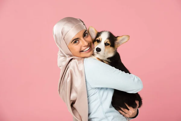 Smiling arabic girl in hijab holding corgi puppy, isolated on pink — Stock Photo