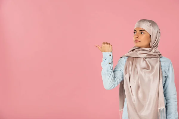 Muslim girl in hijab showing something with thumb, isolated on pink — Stock Photo