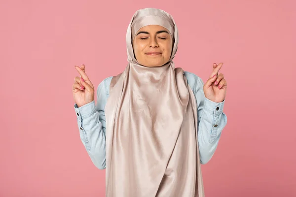 Chica árabe feliz en hijab con los dedos cruzados y los ojos cerrados, aislado en rosa - foto de stock