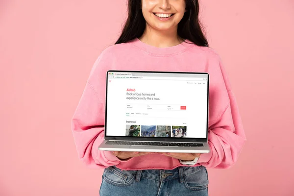 KYIV, UKRAINE - 30 JUILLET 2019 : vue recadrée d'une jeune fille souriante tenant un ordinateur portable avec le site Web airbnb à l'écran, isolée sur rose — Photo de stock