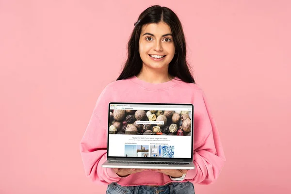 QUIIV, UCRÂNIA - JULHO 30, 2019: menina sorridente segurando laptop com site de fotos de depósito na tela, isolado em rosa — Fotografia de Stock