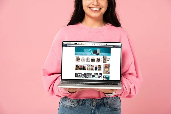 KYIV, UKRAINE - 30 JUILLET 2019 : vue recadrée d'une jeune fille souriante tenant un ordinateur portable avec site web amazone à l'écran, isolée sur rose — Photo de stock