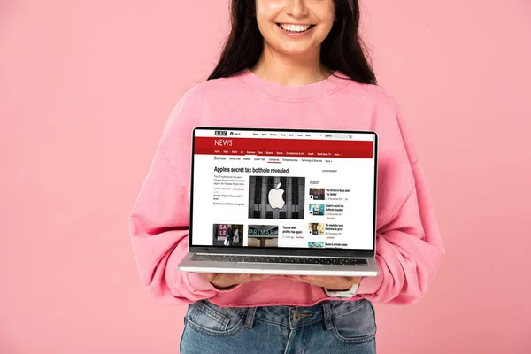 KYIV, UKRAINE - 30 JUILLET 2019 : vue recadrée d'une jeune fille souriante tenant un ordinateur portable avec le site d'information de la BBC à l'écran, isolée sur rose — Photo de stock