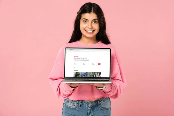 KYIV, UKRAINE - JULY 30, 2019: smiling girl holding laptop with airbnb website on screen, isolated on pink — Stock Photo