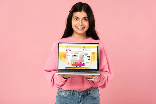 КИЕВ, Украина - 30 июля 2019 года: брюнетка улыбается девушка с ноутбуком с Aliexpress сайт на экране, изолированные на розовый — стоковое фото