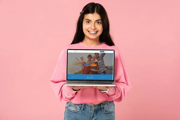KYIV, UKRAINE - JULY 30, 2019: smiling girl holding laptop with couchsurfing website on screen, isolated on pink — Stock Photo