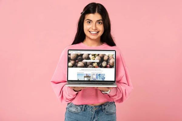 KYIV, UKRAINE - JULY 30, 2019: smiling girl holding laptop with depositphotos website on screen, isolated on pink — Stock Photo