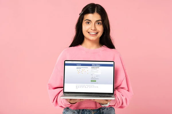 KYIV, UCRANIA - 30 de julio de 2019: niña sonriente sosteniendo el ordenador portátil con el sitio web de facebook en la pantalla, aislado en rosa - foto de stock