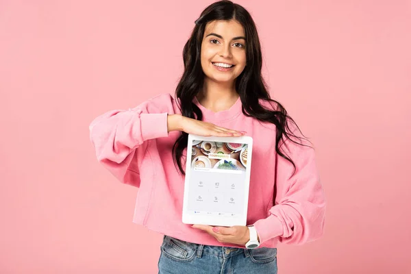 КИЕВ, УКРАИНА - 30 ИЮЛЯ 2019 года: улыбающаяся девушка с цифровым планшетом с четырехквадратным приложением на экране, изолированная на розовом — стоковое фото