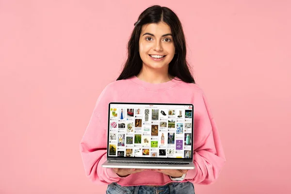 KYIV, UCRANIA - 30 de julio de 2019: niña sonriente sosteniendo el ordenador portátil con el sitio web de pinterest en la pantalla, aislado en rosa - foto de stock