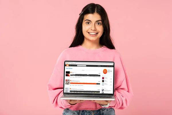 KYIV, UCRAINA - 30 LUGLIO 2019: ragazza sorridente che tiene il computer portatile con il sito web di soundcloud sullo schermo, isolata sul rosa — Foto stock