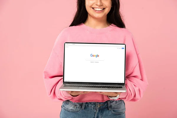 KYIV, UKRAINE - 30 JUILLET 2019 : vue recadrée d'une jeune fille souriante tenant un ordinateur portable avec un site Web google à l'écran, isolée sur rose — Photo de stock