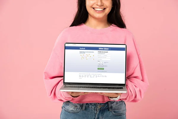 КИЕВ, УКРАИНА - 30 июля 2019 года: обрезанный вид улыбающейся девушки с ноутбуком с facebook сайт на экране, изолированный на розовый — стоковое фото