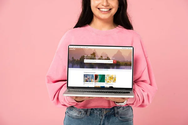 KYIV, UKRAINE - 30 JUILLET 2019 : vue recadrée d'une jeune fille souriante tenant un ordinateur portable avec le site Web shutterstock à l'écran, isolée sur rose — Photo de stock