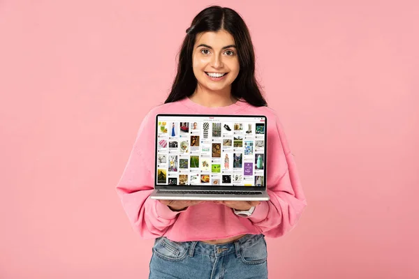 KYIV, UCRANIA - 30 de julio de 2019: niña sonriente sosteniendo el ordenador portátil con el sitio web de pinterest en la pantalla, aislado en rosa - foto de stock