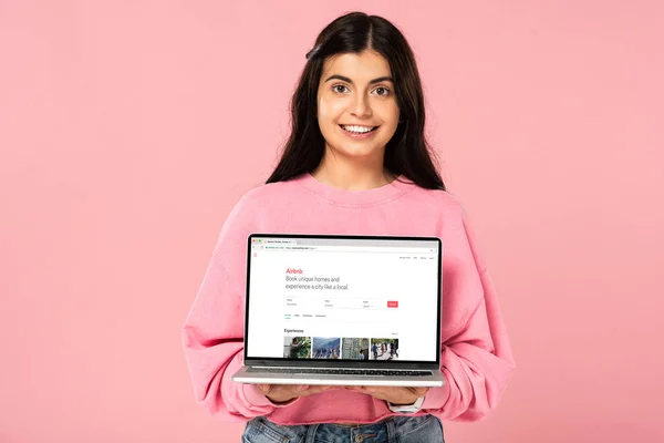 КИЕВ, УКРАИНА - 30 ИЮЛЯ 2019 г.: улыбающаяся девушка с ноутбуком и веб-сайтом airbnb на экране, изолированная на розовом — стоковое фото