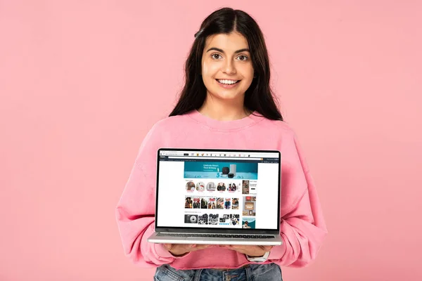 KYIV, UKRAINE - JULY 30, 2019: smiling girl holding laptop with amazon website on screen, isolated on pink — Stock Photo