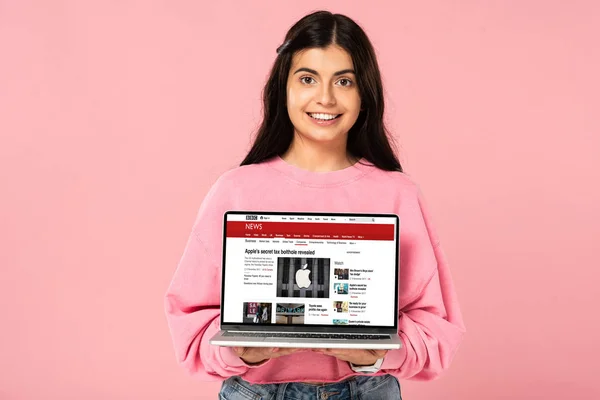 KYIV, UKRAINE - 30 JUILLET 2019 : fille souriante tenant un ordinateur portable avec le site d'information de la BBC à l'écran, isolée sur rose — Photo de stock