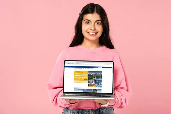 KYIV, UKRAINE - JULY 30, 2019: smiling girl holding laptop with booking website on screen, isolated on pink — Stock Photo