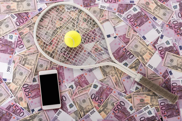 Vista superior del smartphone con pantalla en blanco, raqueta de tenis y pelota en billetes de euro y dólar, concepto de apuestas deportivas - foto de stock