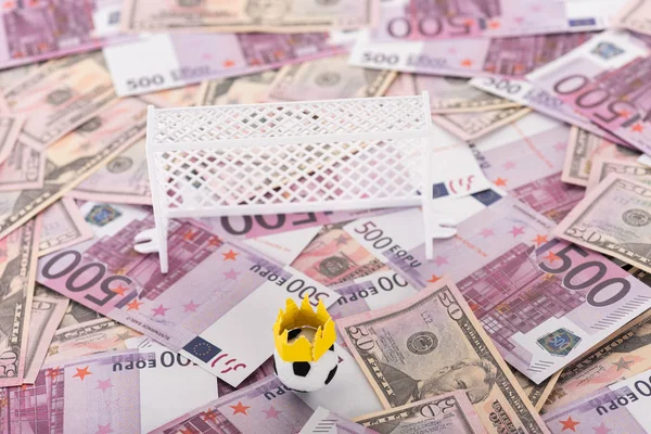 Pallone da calcio giocattolo con corona di carta vicino ai cancelli di calcio in miniatura su banconote in euro e dollaro, concetto di scommesse sportive — Foto stock