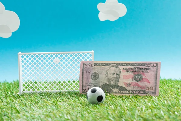 Pelota de fútbol de juguete y puertas cerca de billete de dólar sobre fondo azul con nubes, concepto de apuestas deportivas - foto de stock