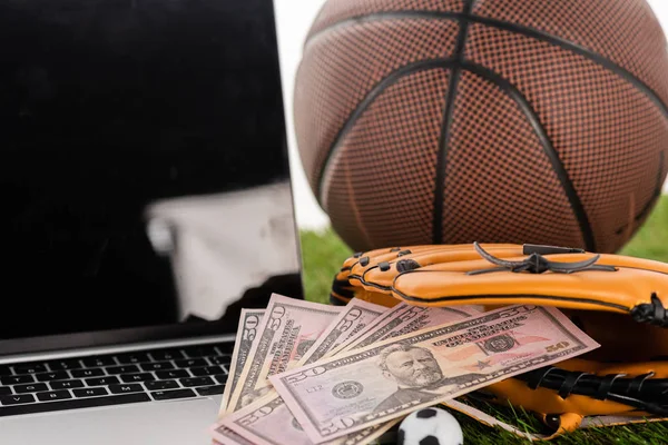 Крупным планом банкнот в бейсбольной перчатке рядом с игрушечным футбольным мячом, ноутбуком и баскетбольным мячом, изолированным на белом, концепция ставок на спорт — стоковое фото