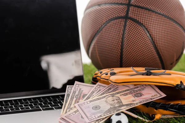 Vista da vicino delle banconote da un dollaro nel guanto da baseball vicino a pallone da calcio giocattolo, laptop e pallone da basket isolati su bianco, concetto di scommesse sportive — Foto stock