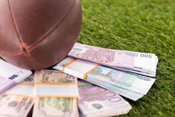 Enfoque selectivo de paquetes de billetes de euro y dólar cerca de la pelota de rugby en hierba verde, concepto de apuestas deportivas - foto de stock