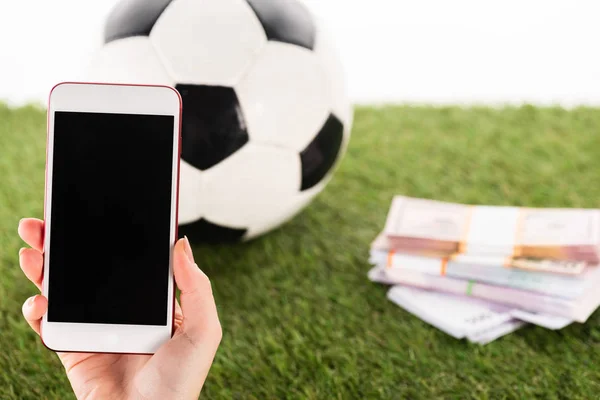 Vista parcial de la mano femenina con smartphone cerca de paquetes de dinero y pelota de fútbol sobre hierba verde aislado en blanco, concepto de apuestas deportivas - foto de stock