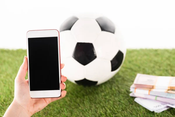 Vista recortada de la mano femenina con teléfono inteligente cerca de paquetes de dinero y pelota de fútbol sobre hierba verde aislado en blanco, concepto de apuestas deportivas - foto de stock