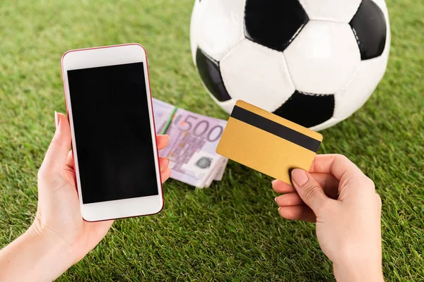 Vista recortada de las manos femeninas con teléfono inteligente y tarjeta de crédito cerca de la pelota de fútbol y billetes en euros en hierba verde, concepto de apuestas deportivas - foto de stock