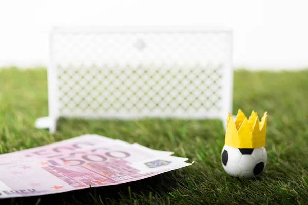 Foco selectivo de pelota de fútbol de juguete con corona de papel cerca de billetes en euros y puertas en miniatura aisladas en blanco, concepto de apuestas deportivas - foto de stock