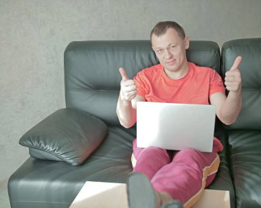 Dizüstü bilgisayarlı genç adam evdeki kanepede oturur ve başparmağını kaldırır, kameraya bakar.