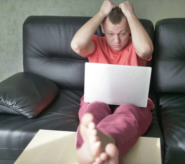 Удивленный молодой человек сидит на диване с ноутбуком в гостиной — стоковое фото