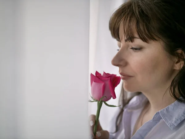 Junge brünette Frau schnuppert eine Rose, die am Fenster steht — Stockfoto