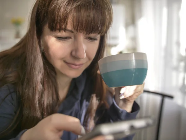 Молодая женщина держит кофейную кружку и с помощью телефона на кухне — стоковое фото