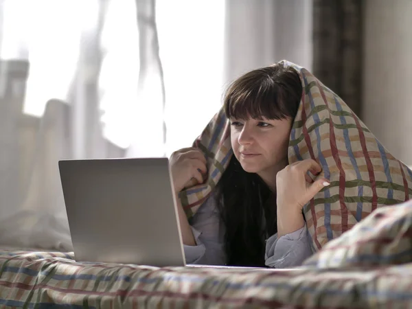 Брюнетка лежит под одеялом и смотрит в ноутбук — стоковое фото