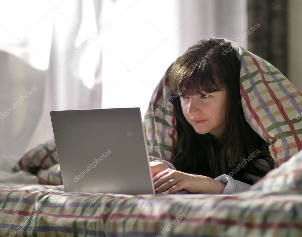 A young brunette woman is lying under a blanket and typing in a laptop.