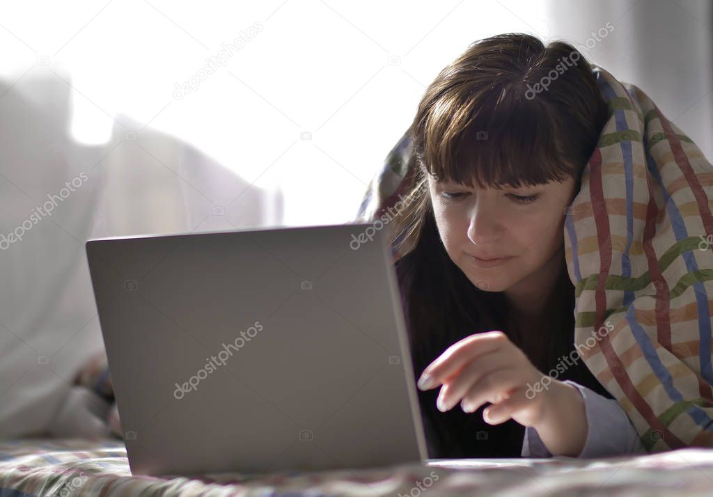 A young brunette woman is lying under a blanket and typing in a laptop.