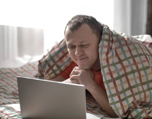 Joven hombre sonriente está acostado en la cama, mirando en el portátil . — Foto de Stock