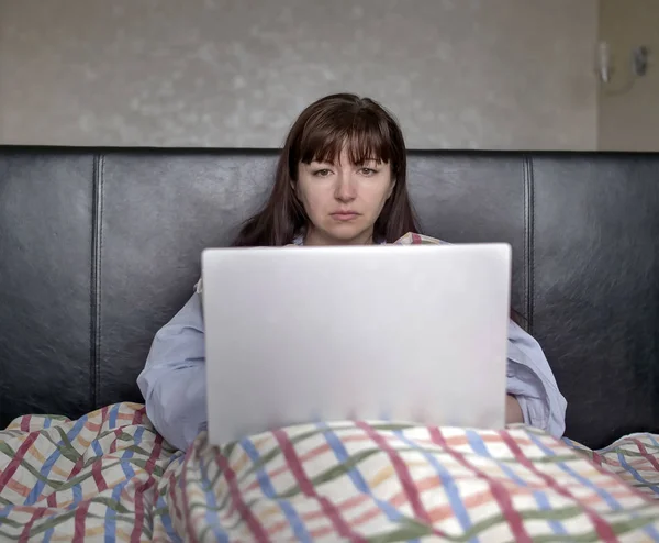 Серьезная молодая женщина сидит в постели под одеялом с ноутбуком — стоковое фото