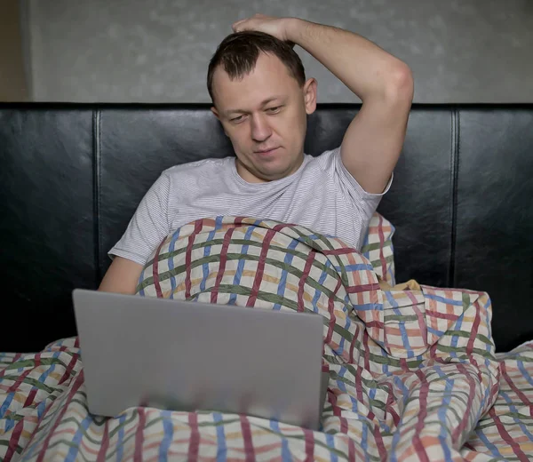 Молодой человек сидит в постели под одеялом с ноутбуком — стоковое фото