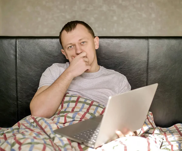 Потрясающий молодой человек, сидящий в кровати с ноутбуком, глядя прямо в камеру — стоковое фото