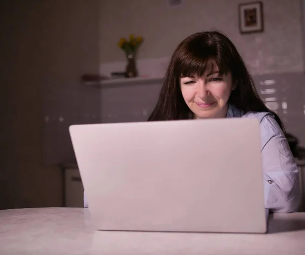 Милая улыбающаяся юная брюнетка, сидящая вечером на кухне с ноутбуком — стоковое фото