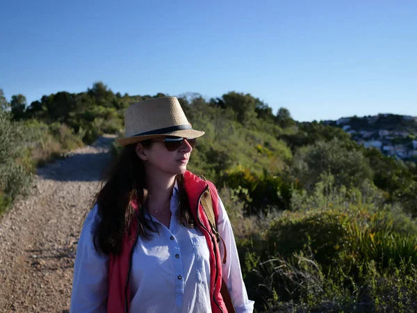 Jolie brune touristique dans un chapeau et des lunettes de soleil va le long du chemin sur la colline — Photo