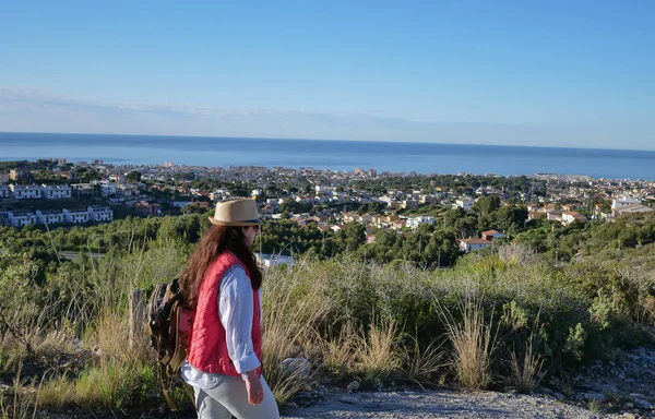 Fille touristique dans un chapeau et un sac à dos va le long du chemin sur une colline à la ville par la mer — Photo