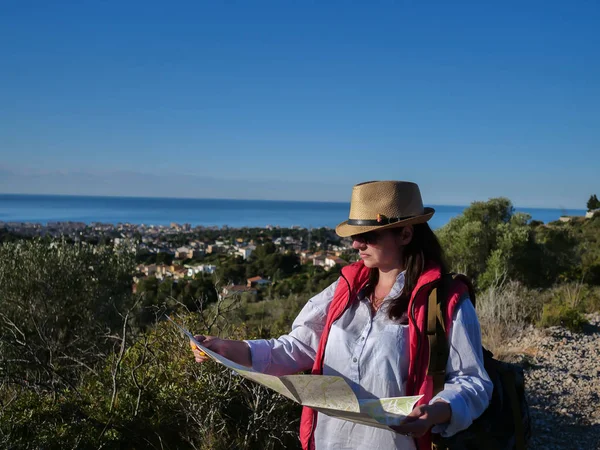 Attractive girl tourist in a hat is considering a map while standing on the trail