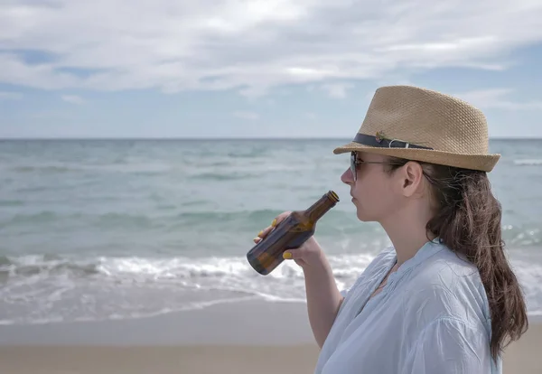 Cute brunette girl in a hat drinks beer standing near the sea — Stock Photo, Image